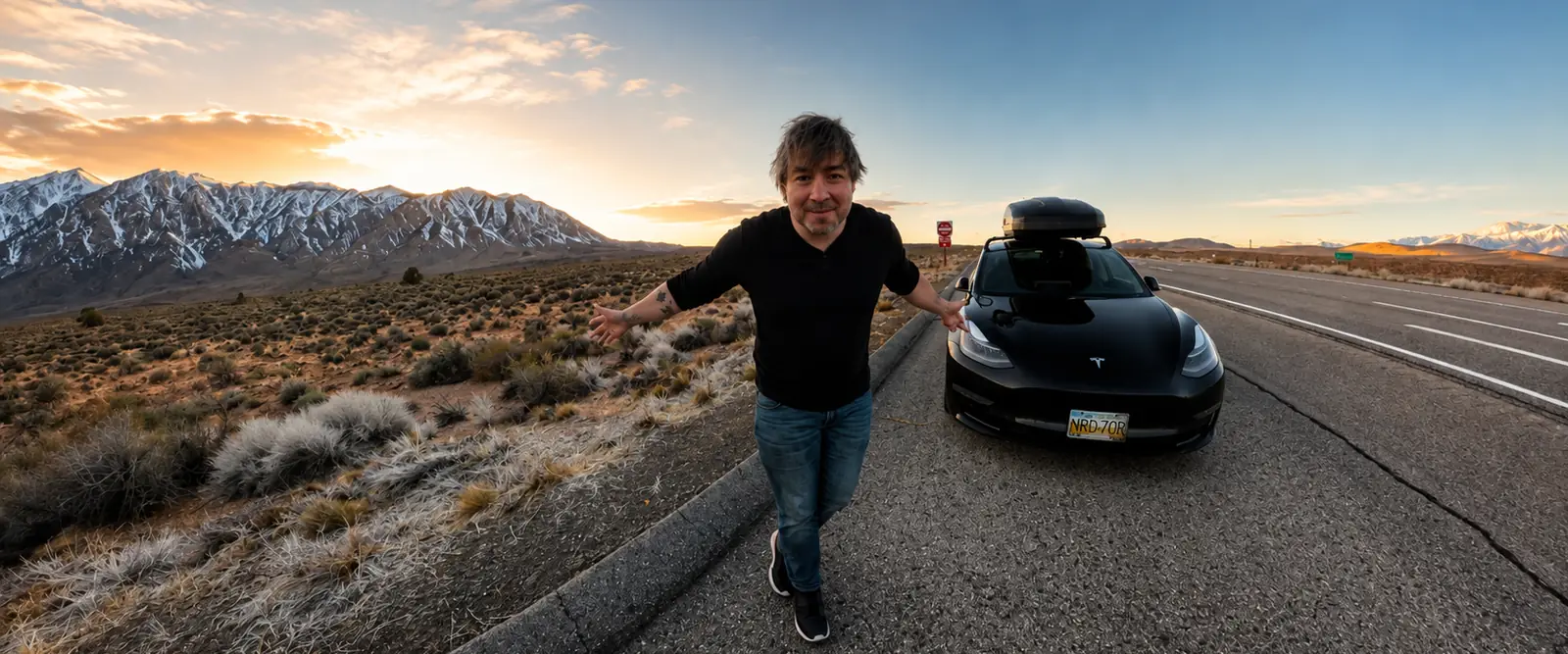 Joey Caughey standing in the middle of a desert highway at sunrise, arms out, beside a black car with a roof box; snow on the mountains behind